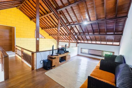 Spacious tropical loft interior with wooden beamed ceiling, yellow brick accent wall, entertainment console, and rattan sofa on hardwood floor. - Bali Villas